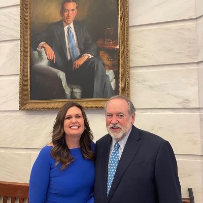 Mike Huckabee is wearing a black suit with the blue tie. Sarah Sanders is wearing a blue dress 